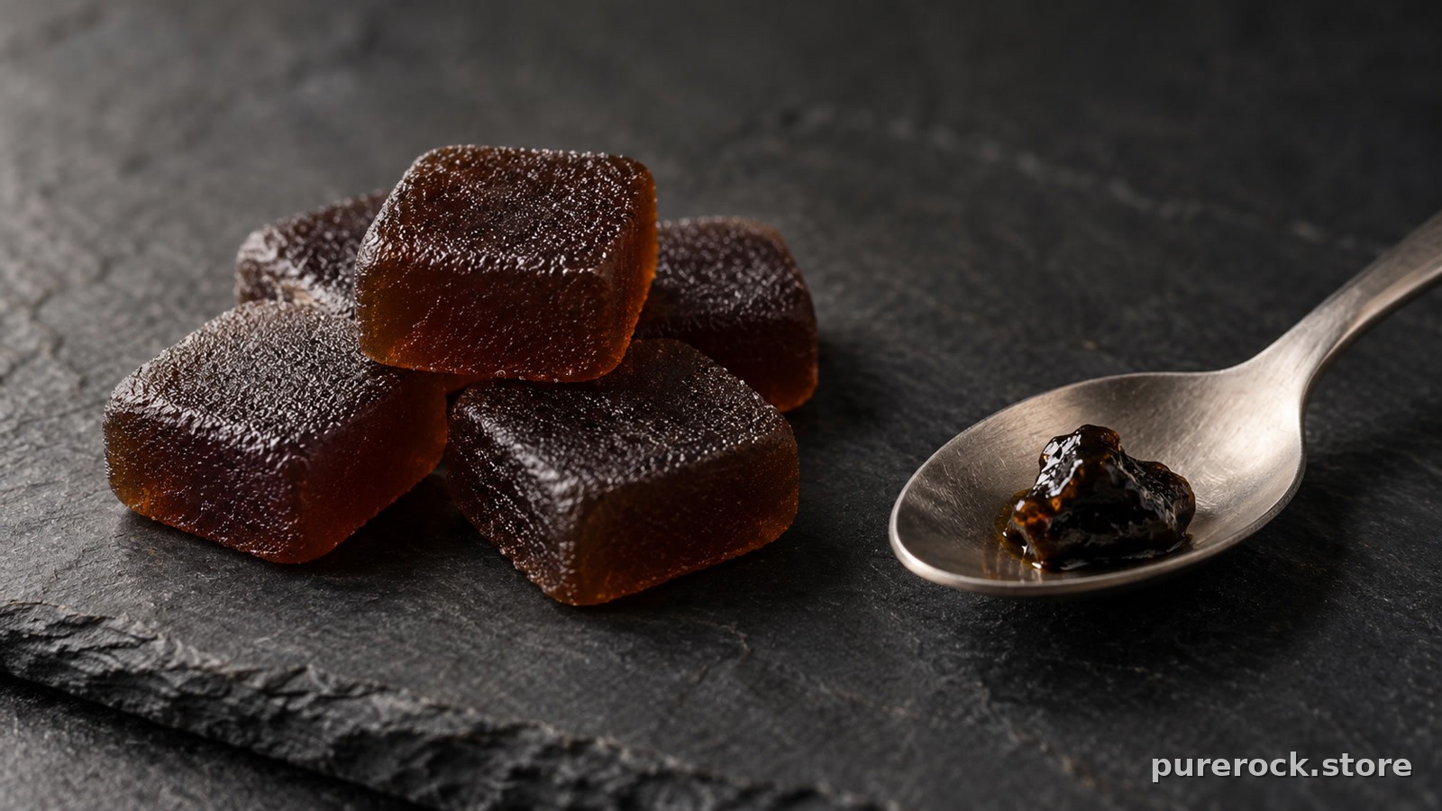 A small pile of dark, unbranded square gummies on a slate surface next to a spoon holding a pea-sized piece of dark resin